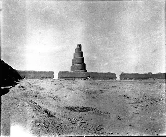 Black-and-white 1909 photograph: the spiral Malwiya minaret of the Great Mosque of Samarra rises behind a ruined mud-brick enclosure wall, bare rubble-strewn ground stretching toward the wall under a pale desert sky.