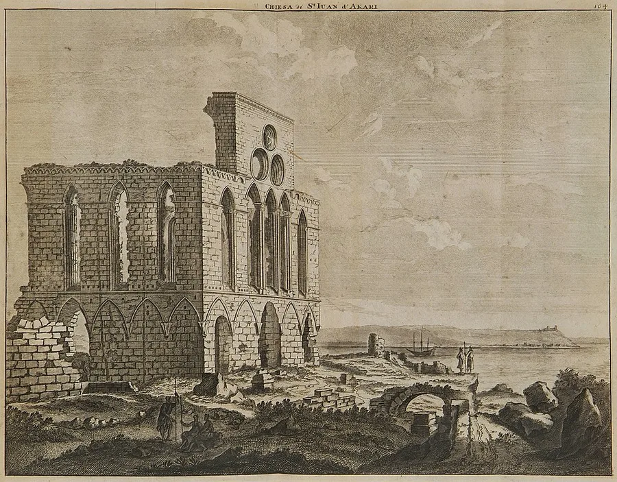 An 18th-century engraving of a roofless ruined Gothic church at Acre: a stone facade with tall arched windows and round oculi, set on a rocky shoreline with a walled point and ships in the bay behind, and small robed and seated figures near the ruin's base.