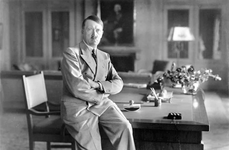 Black-and-white photograph of Adolf Hitler, in a pale double-breasted suit, leaning against a desk with arms folded in a wood-paneled room at the Berghof, mid-1930s.
