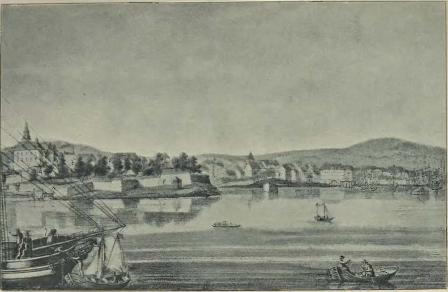 Monochrome-toned harbor view of Christiania (Oslo) from the water: sailing ships and rowboats in the foreground, a low walled fortification on a point of land at center-left, and the town's buildings and hills along the shoreline under an overcast sky.