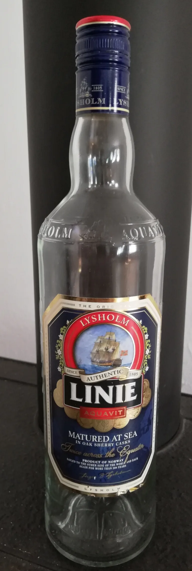 Snapshot of a clear Lysholm Linie Aquavit bottle, navy cap, leaning against a tall dark gift canister on carpet at home; the label shows a sailing ship and reads 'Authentic Linie Aquavit, Matured at Sea in Oak Sherry Casks, Twice across the Equator, Product of Norway.'