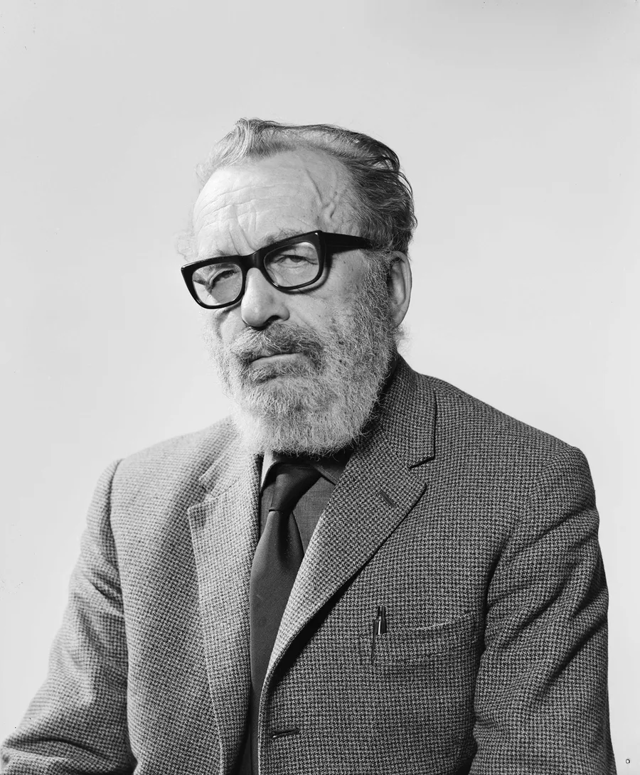 Black-and-white 1963 studio portrait of an older man with a full grey beard, heavy dark-framed glasses, and swept-back hair, wearing a tweed jacket and tie, photographed from the chest up against a plain light background.
