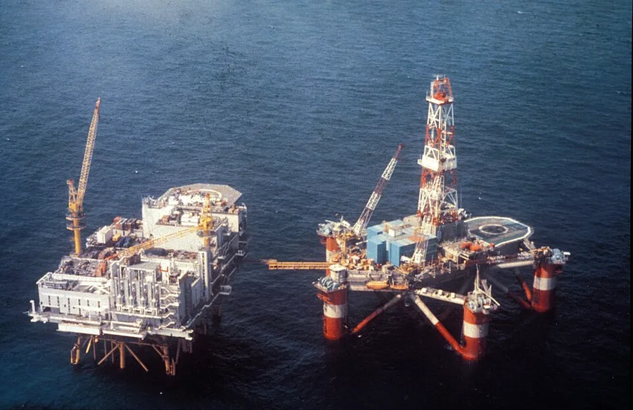Aerial photo of two offshore platforms in the North Sea before 1980: a boxy production platform on the left and a semi-submersible rig with a red-and-white derrick on the right, moored close together on calm water.