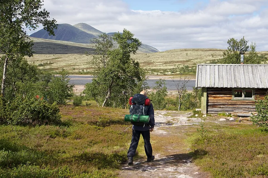 A hiker with a loaded backpack and rolled sleeping mat walks away from the camera along a path through open, unfenced Norwegian mountain heath toward a small timber hut, with birch scrub, a lake, and bare fells in the background.