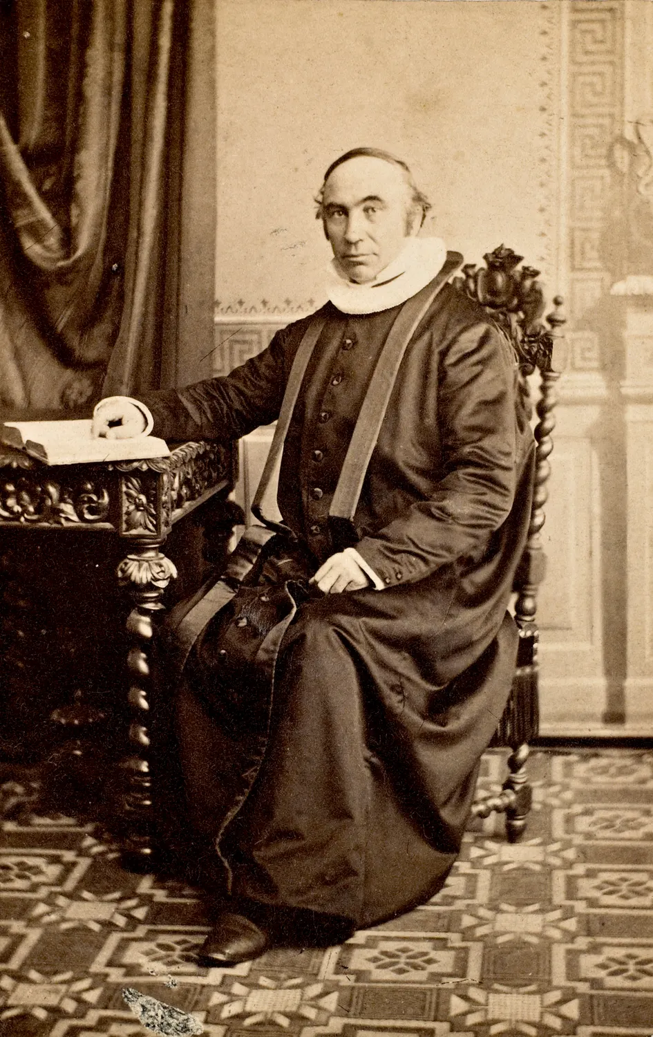 Jørgen Moe seated in the dark cassock and white ruff of a Norwegian priest, one hand resting on an open book — a carte-de-visite from the Olsen & Thomsen studio, Christiania, c. 1865.
