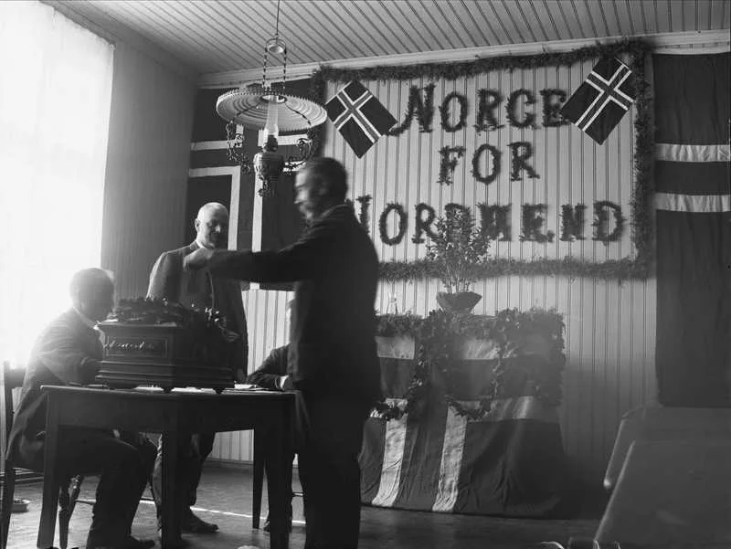 Black-and-white photograph of a room decorated with Norwegian flags and a banner reading 'Norge for Nordmænd,' a ballot box on a table, and men gathered around it — a decorated polling place during the 13 August 1905 referendum.