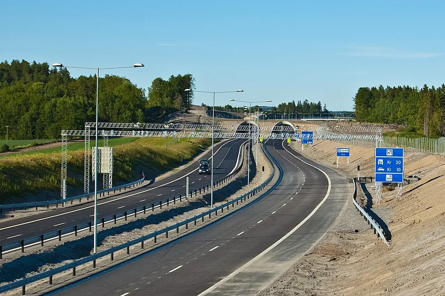 An E18 highway near Larvik with an overhead lattice-truss gantry carrying toll-reading equipment across the carriageway, blue signs reading 'Kr 20' and 'Kr 10' beside the road, no barrier, booth, or stop in sight.