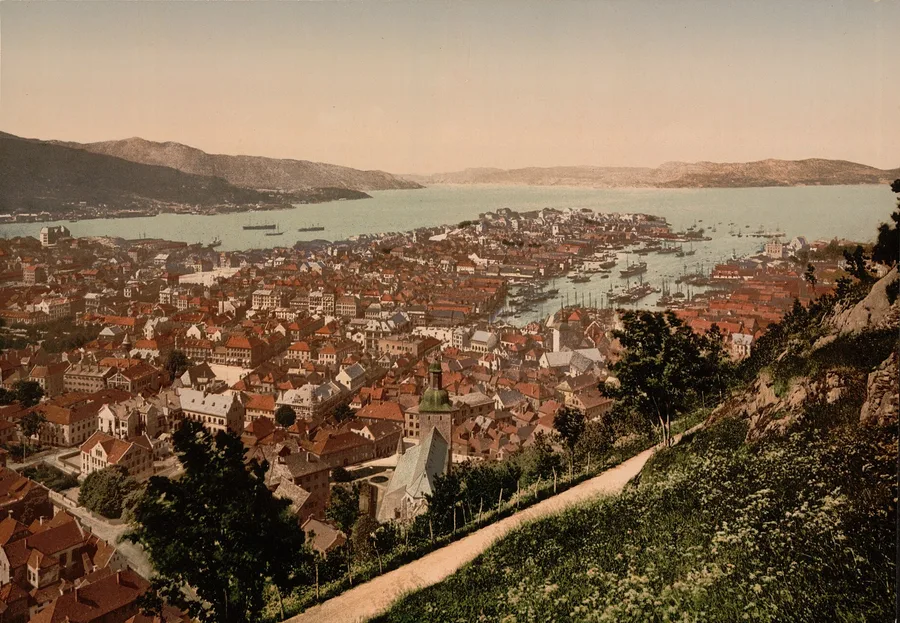 A colored 1890s photochrom of Bergen from the slope of Fløyfjellet: red rooftops and a green-spired church tower below a hillside path, Vågen harbour dense with masted ships, and the Byfjorden opening between low mountains to the horizon.