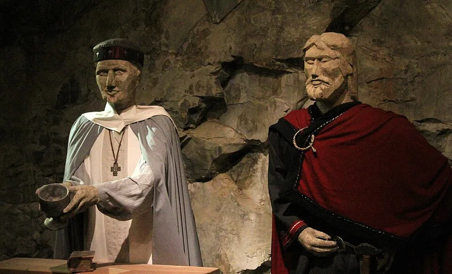 Carved wooden figures of Bishop Grimkjell (grey cape, dark cap, holding a chalice) and King Olav (red-and-black robe with a round brooch) in a staged museum tableau at Moster Amfi, lit against a dim stone-cave backdrop.