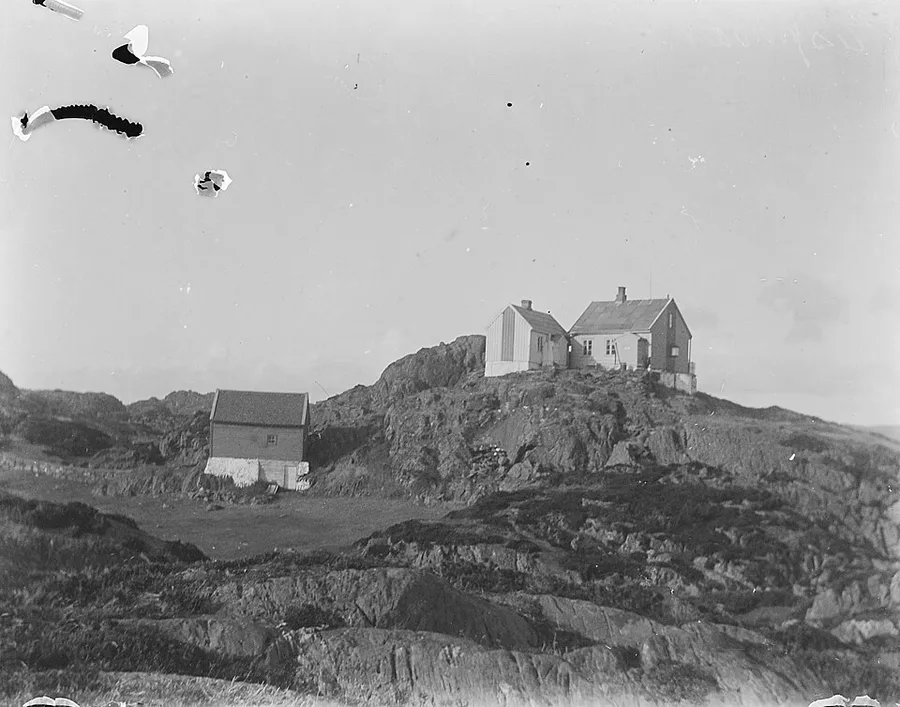 Late-19th-century black-and-white photograph of a few wooden houses perched on a rocky, grass-tufted hilltop at Espevær, Bømlo, under a wide pale sky with scattered old photo-emulsion damage marks.