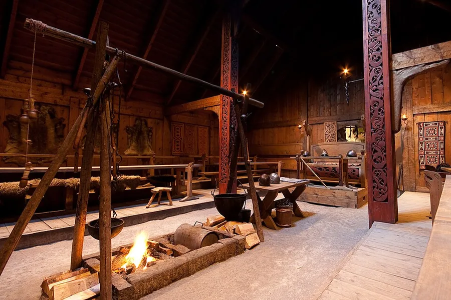 Interior of the reconstructed chieftain's longhouse at Borg in Lofoten: a fire burns in the open hearth beneath a cauldron slung from a timber crane, with carved red-painted posts, fur-draped benches, woven wall hangings and the dim timber roof receding down the hall.