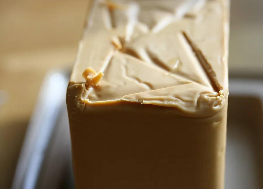 Extreme close-up of a caramel-brown block of brunost cheese, its glossy cut top surface catching the light, sitting on a wooden counter.
