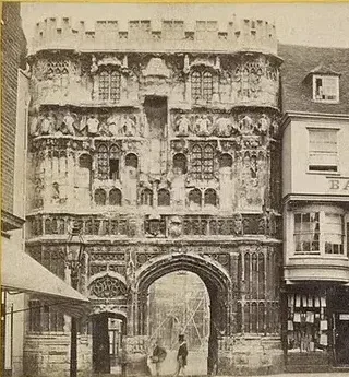 Sepia photograph of the ornately carved stone Christ Church Gate, the main entrance to Canterbury Cathedral's precincts, with tiers of heraldic panels, corner turrets, and pedestrians passing through the archway below.