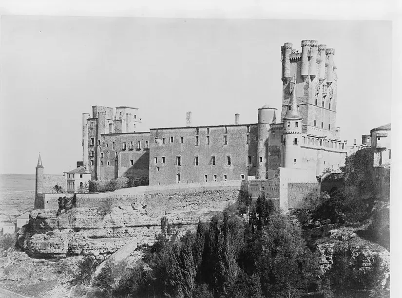 Albumen photograph of the Alcázar of Segovia massed on its rock spur above a wooded gorge — long fortress walls and round turrets, the tall keep of Juan II with clustered turrets at right, and the bare Castilian meseta stretching to the horizon at left.