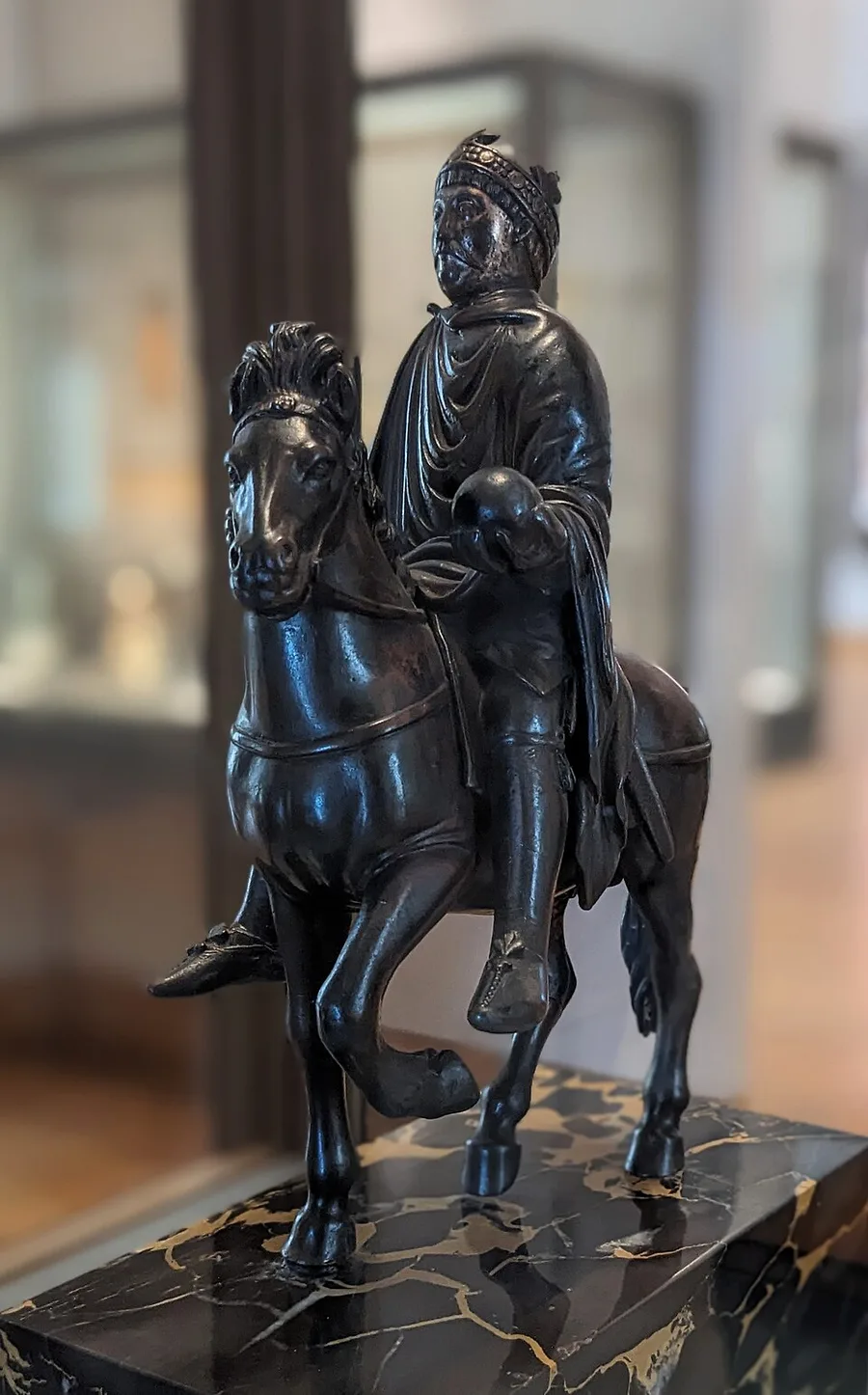 A dark bronze statuette of a crowned, moustached rider in a draped cloak holding an orb in his outstretched hand, seated on a pacing horse, displayed on a gold-veined black marble plinth against a blurred museum gallery.