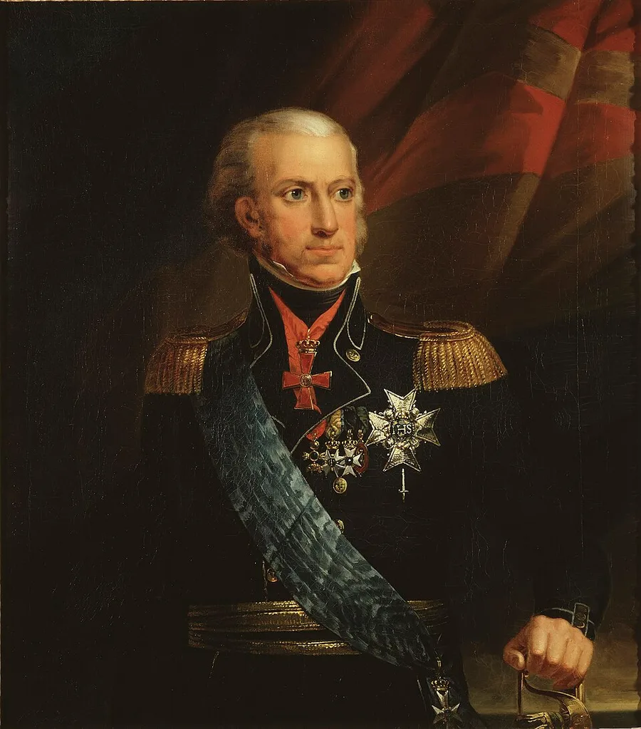 Formal half-length portrait of an elderly king with white hair and a receding hairline, standing in a dark military coat hung with medals and a blue sash, one hand resting at lower right, a red flag bearing a cross behind him.