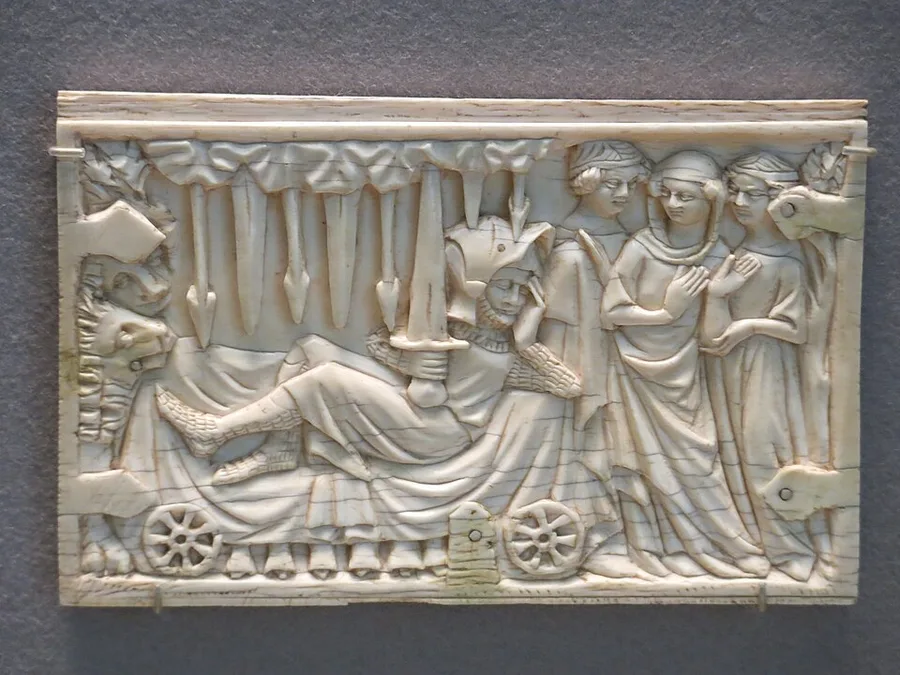 A carved ivory casket panel showing a knight reclining on a wheeled bed clutching a sword, a lion crouched at its foot, while a group of women look on — the 'Gawain in the Perilous Bed' scene from Perceval.