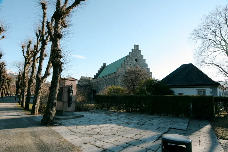 The Christ Church site at Bergenhus in Bergen: a flagstone-paved plot edged by low hedges with a small dark memorial statue on a stone plinth, pollarded trees lining the path at left, and the stepped stone gable and green roof of Håkon's Hall rising behind under a clear sky.