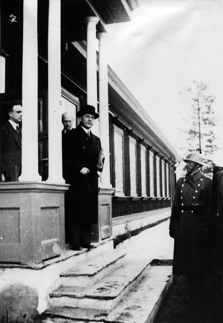 Black-and-white 1940 photograph of three men in dark coats and hats on the covered porch of a wooden building at Elverum, a Norwegian soldier in a steel helmet standing in the foreground.