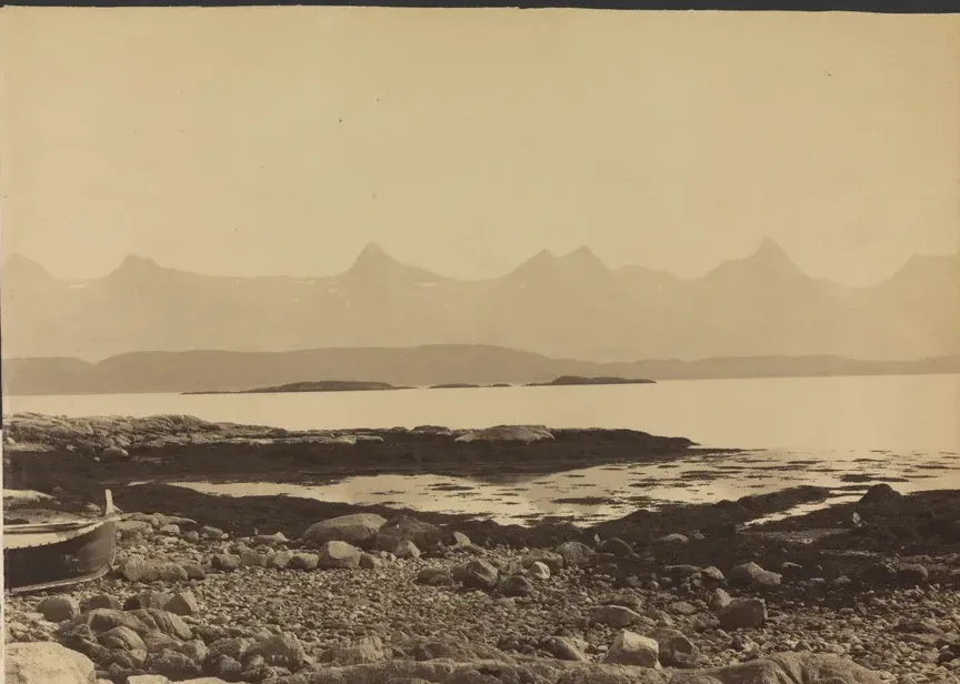 Knud Knudsen's nineteenth-century photograph of De syv søstre — the seven peaks rising in a row across the water behind a rocky foreshore and the bow of a beached boat.