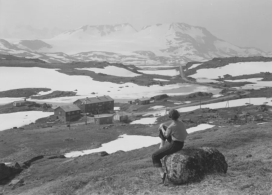A 1957 black-and-white photo of Sognefjellshytta, a cluster of mountain-lodge buildings on open, snow-patched fell terrain, with a young visitor seated on a boulder in the foreground looking toward snow-capped peaks.