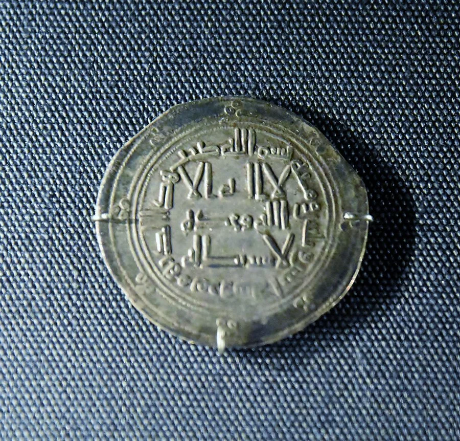 A silver Abbasid dirham pinned to dark blue display fabric, one face filled edge to edge with lines of Kufic Arabic script ringed by a circular marginal inscription — no portrait or image, only lettering.