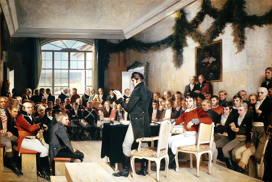 Oscar Wergeland's 1885 history painting of the crowded Eidsvoll assembly hall: a standing delegate mid-oration at a draped table, chairs and dozens of formally dressed men seated and standing around him, a portrait on the back wall.