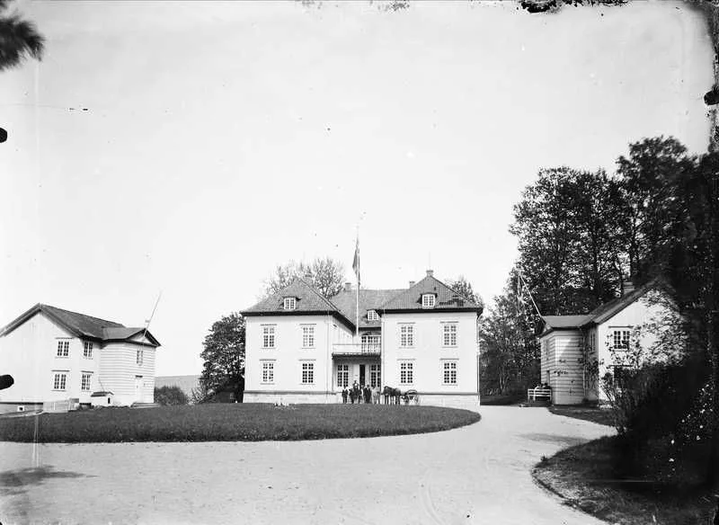 Black-and-white photograph, circa 1890, of Eidsvollsbygningen: a two-story white manor house with a flagpole, flanked by outbuildings, seen across a gravel forecourt with a horse and carriage.