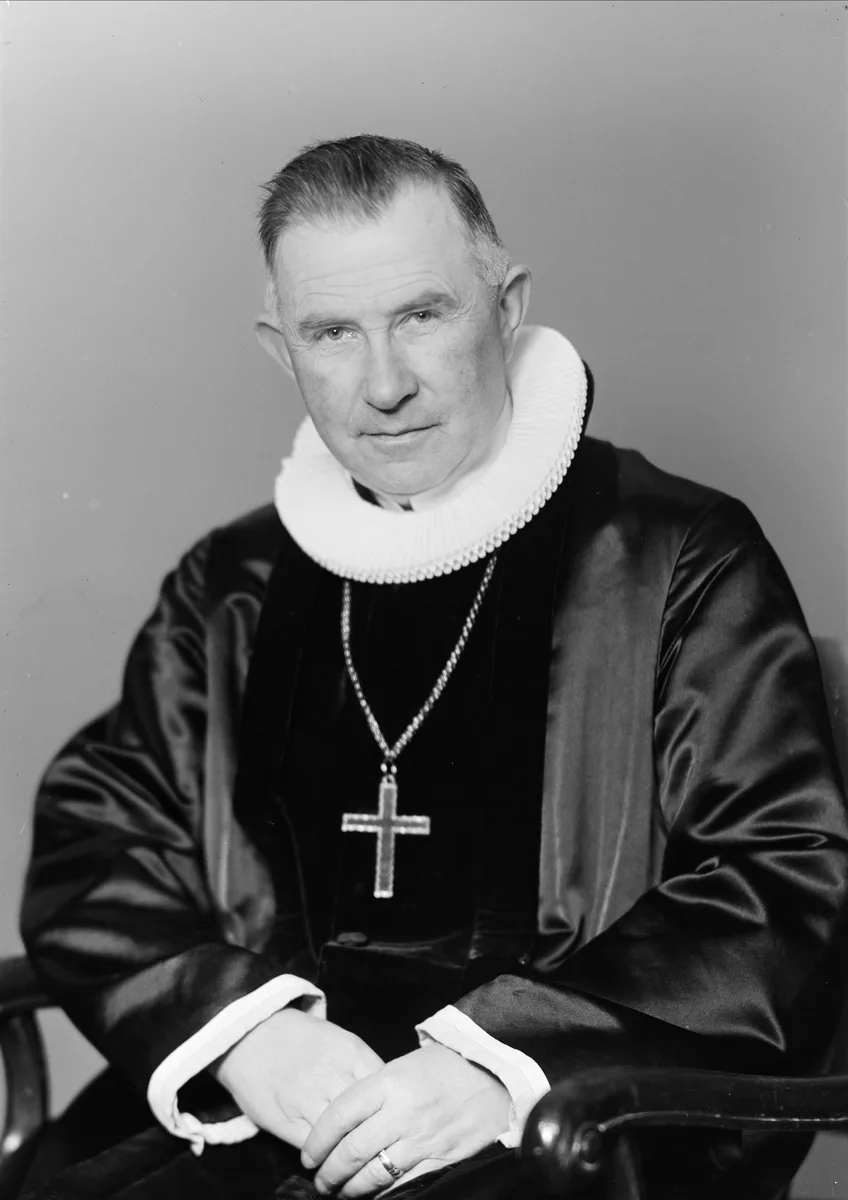 Black-and-white studio portrait, half-length, of an older man in clerical robes and a wide pleated ruff collar, a pectoral cross on a chain around his neck, looking directly at the camera.