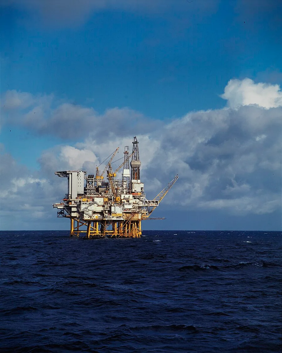 A single steel offshore oil platform standing in the North Sea, its derrick, cranes, and helideck rising above a yellow-legged jacket structure, against a partly cloudy sky.
