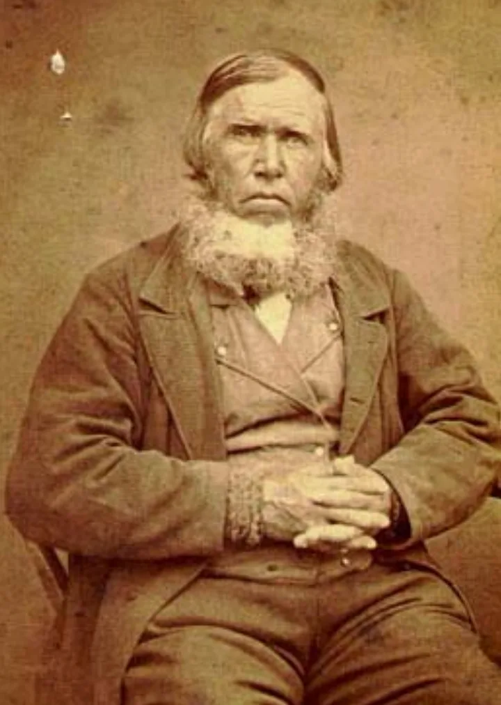 Sepia studio photograph of an elderly bearded man in a dark suit coat and waistcoat, hair combed back, seated with hands clasped in his lap, looking directly at the camera against a plain backdrop.