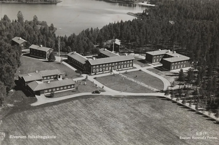 Aerial black-and-white photograph of the Elverum folk high school, a cluster of large wooden buildings around an open lawn beside a lake, ringed by forest.