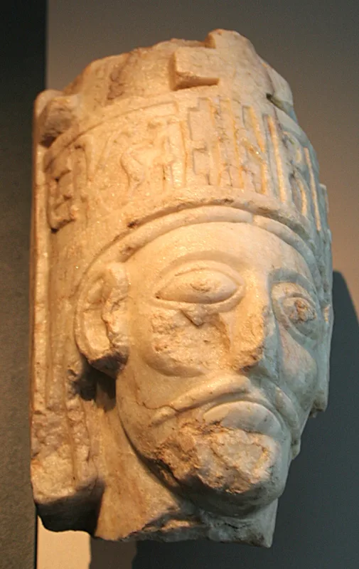 A weathered marble bust of a crowned king, carved in relief with a banded crown showing faint carved figures and worn lettering, photographed at an angle against a plain gray backdrop.