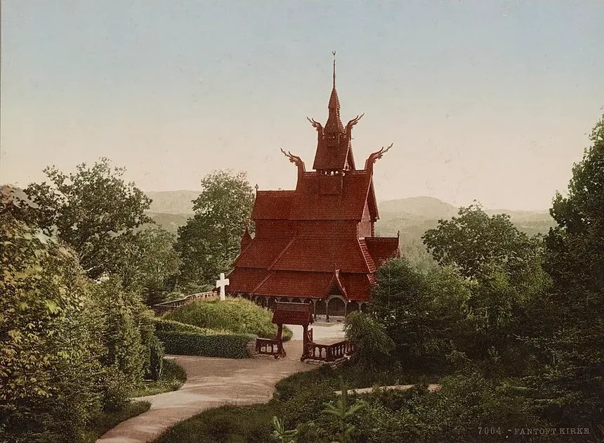 A hand-tinted photochrom print of the original Fantoft Stave Church on its wooded Bergen hillside, dragon finials and a tiered dark-red shingled roof, with a white memorial cross and a gated path in the foreground, decades before the 1992 arson.