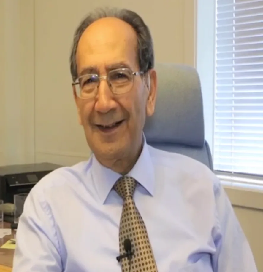 Head-and-shoulders photo of Farouk Al-Kasim, an older man with grey hair and glasses, smiling, seated in an office in front of a window with blinds.