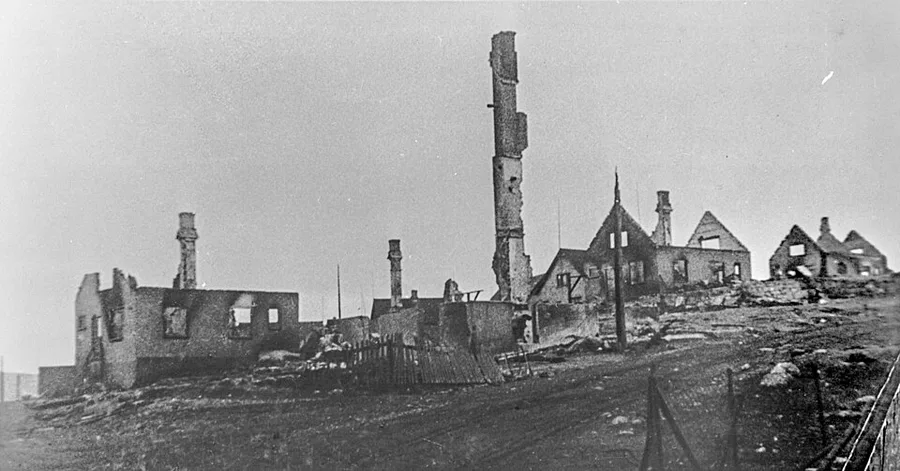 Black-and-white photograph of burned-out building ruins in Kirkenes, roofless brick walls and several standing chimneys amid rubble after the 1944 scorched-earth destruction.