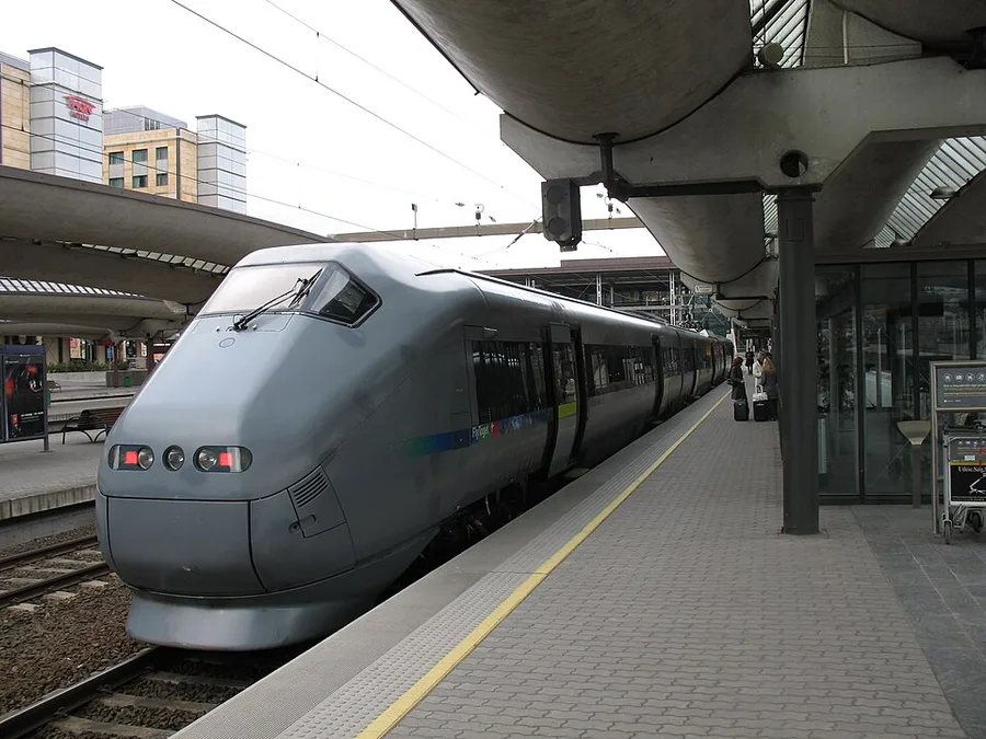 A silver-grey Flytoget airport express train, its sleek aerodynamic nose facing the camera, stands at an open-air platform beneath Oslo Central Station's curved concrete canopies; a thin blue-green stripe runs along its side and travelers with luggage wait on the platform.