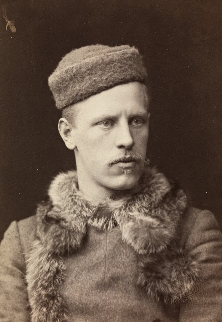Sepia 1888 studio portrait of a young, mustached Fridtjof Nansen in a fur cap and thick fur-collared coat, head turned slightly to the side against a dark backdrop.