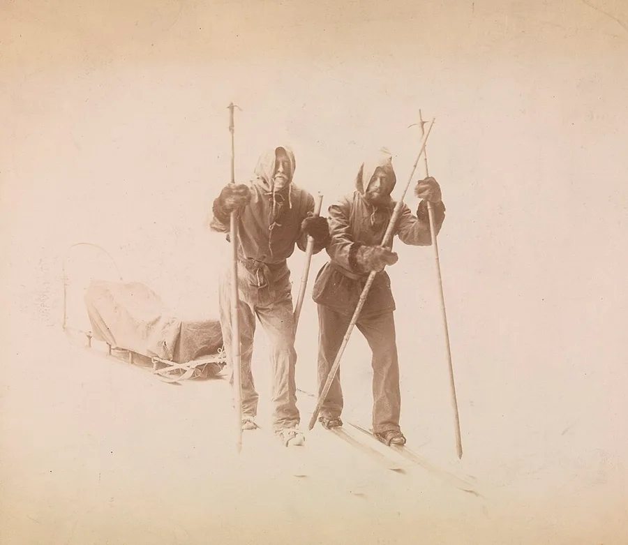 Sepia studio photograph of two bearded men in hooded polar travel clothing and fur mittens, standing on skis with ski poles planted in the snow, a loaded sledge behind them, against a plain misty backdrop.