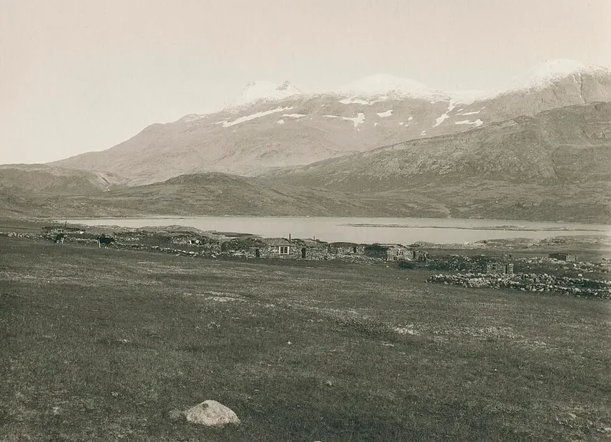 Sepia photograph across a grassy plain at Igaliku: low stone-built houses and drystone walls of the small village on the old bishopric ground, with the calm fjord behind and snow-capped mountains rising above.