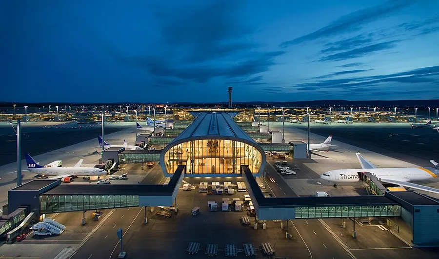 Aerial night view of Oslo Airport Gardermoen's curved glass-and-timber terminal glowing with interior light, control tower rising behind it, and SAS, Norwegian, and Thomas Cook aircraft parked at jet bridges on the lit tarmac.