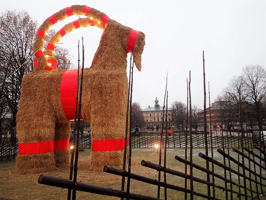 The giant straw Gävle goat standing ribboned and lit on Slottstorget in December 2019, its protective fence in front and the town hall behind.