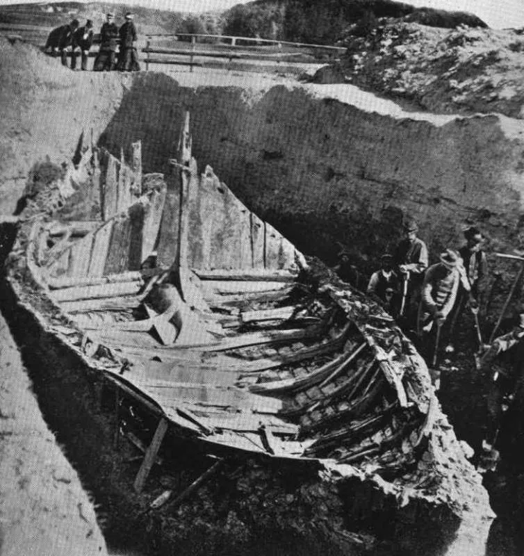 Halftone 1880 photograph of the Gokstad excavation: the ship's timber hull with exposed ribs lies in a deep trench cut through the burial mound, broken uprights rising amidships, workmen with tools at right and onlookers lining the mound's edge above.
