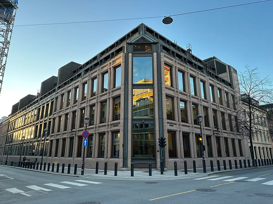 Corner view of the modern Norges Bank headquarters in Oslo at dawn — a massive granite-clad block with deep-set window grids, a glass corner bay, and bollard-lined sidewalks along two empty streets.
