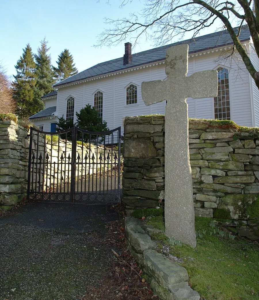 A weathered stone cross stands beside the wrought-iron gate and stone wall at the entrance to Eivindvik church on Gulen, the white clapboard church with pointed Gothic windows visible behind it among evergreens.