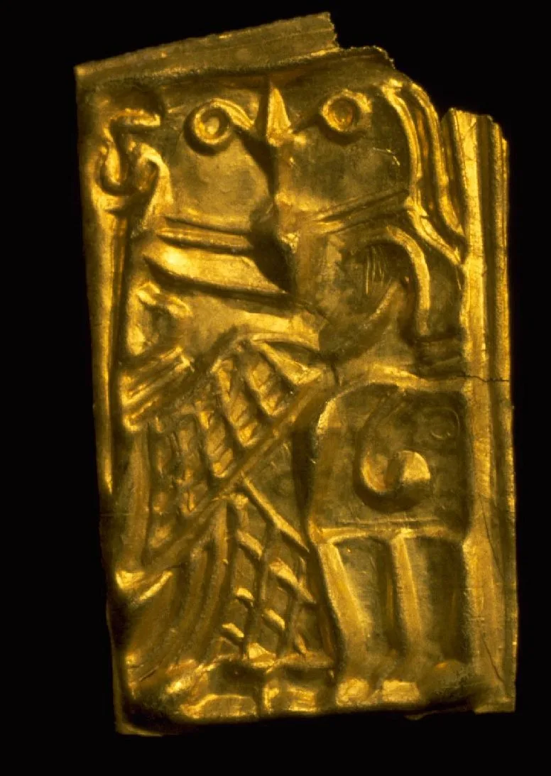 Macro photograph against black of a small rectangular stamped gold-foil gullgubbe, no larger than a fingernail, embossed with a man and a woman in profile, heads close together cheek to cheek.