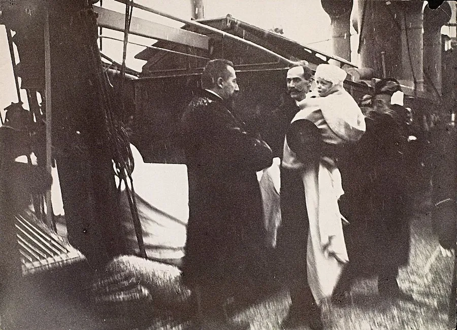 Haakon VII, in a dark naval greatcoat, carries the infant Crown Prince Olav bundled in white across a ship's deck on 25 November 1905, while Prime Minister Michelsen stands facing him at left amid the rigging.