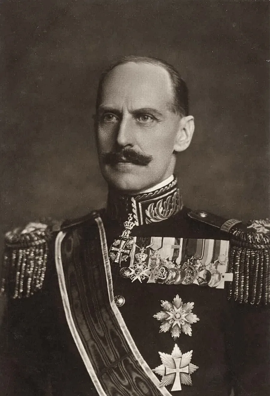 Sepia-toned studio portrait of King Haakon VII of Norway in a military dress uniform with fringed epaulettes, an array of medals, and a wide ceremonial sash, looking off to the side.