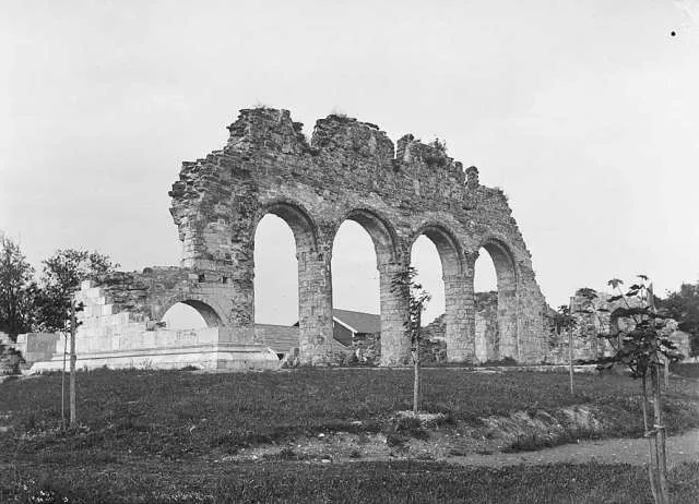 A sepia 19th-century photograph of the Hamar cathedral ruins: four roofless stone arches and adjoining walls standing in an open grassy field, young trees planted along the foreground.