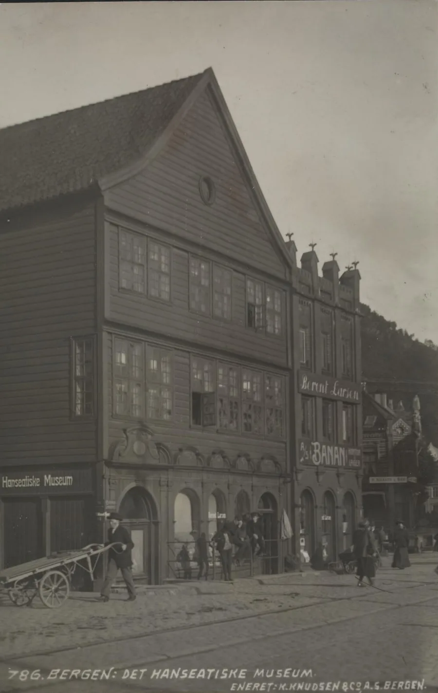 K. Knudsen & Co. postcard photograph: the dark timber gable of Finnegården fills the frame head-on, a 'Hanseatiske Museum' sign over its street-level corner, a hand-cart and passers-by on the cobbles, with the caption '786. Bergen: Det Hanseatiske Museum' below.